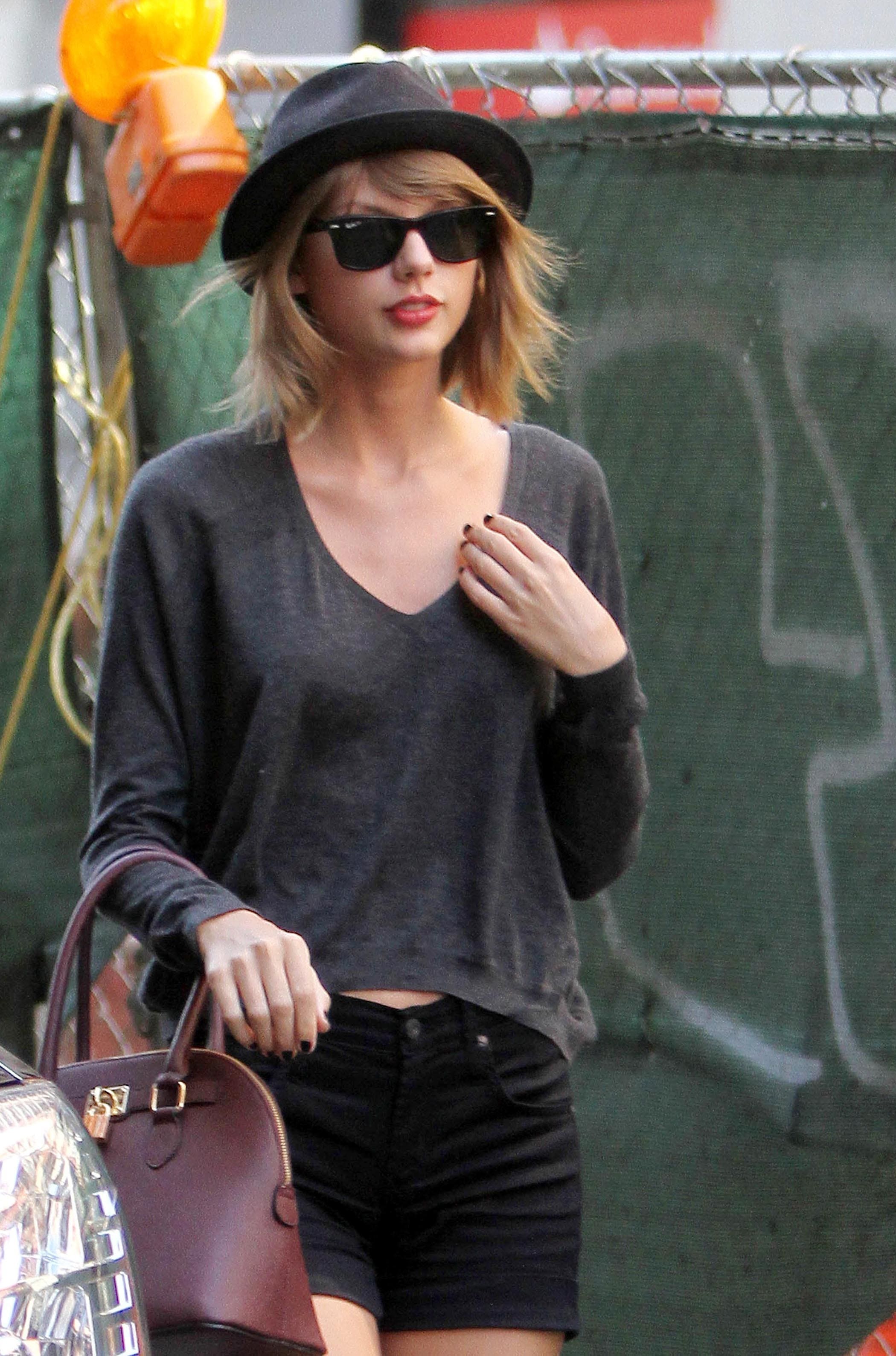 Taylor Swift in cropped gray sweater and black shorts with hat
