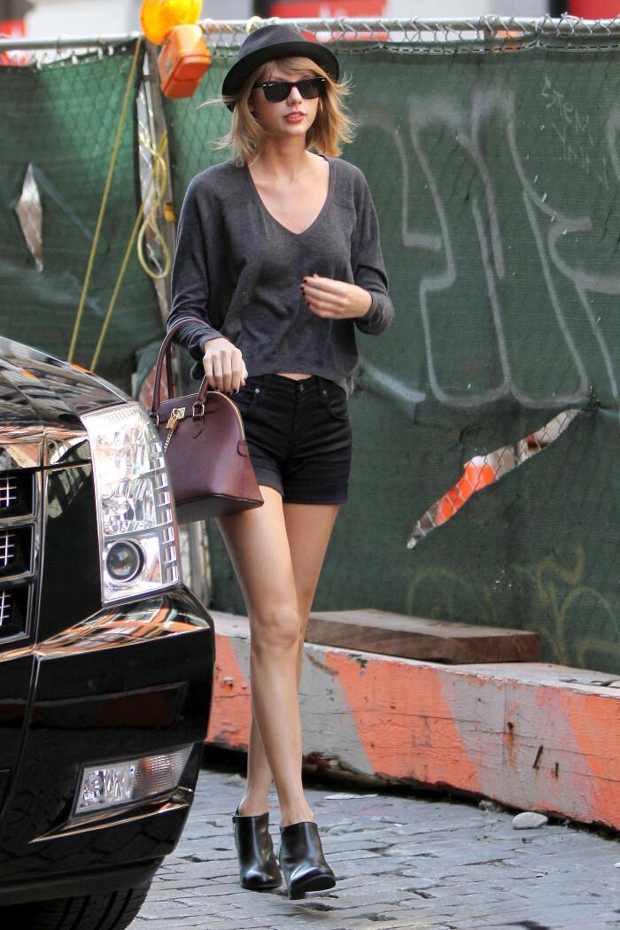 Taylor Swift in gray V-neck sweater, black shorts, boots, and hat